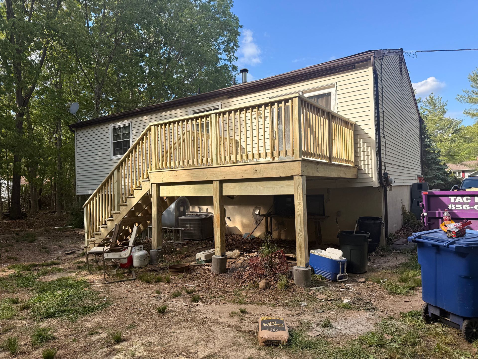House with raised wooden deck and stairs, viewed from a yard with trees and trash bins.