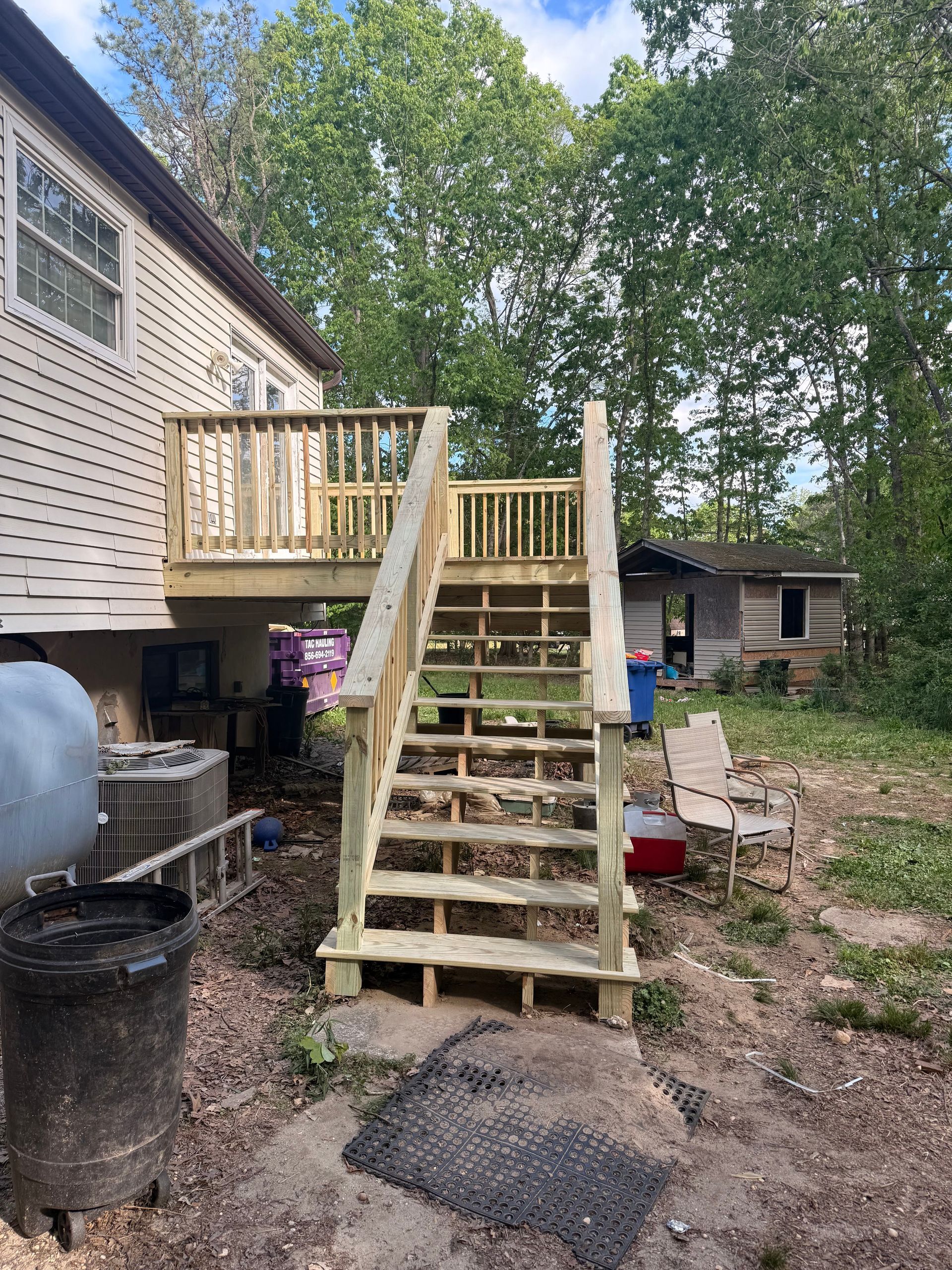 Back deck with stairs leading to a yard beside a house, surrounded by trees and outdoor items.