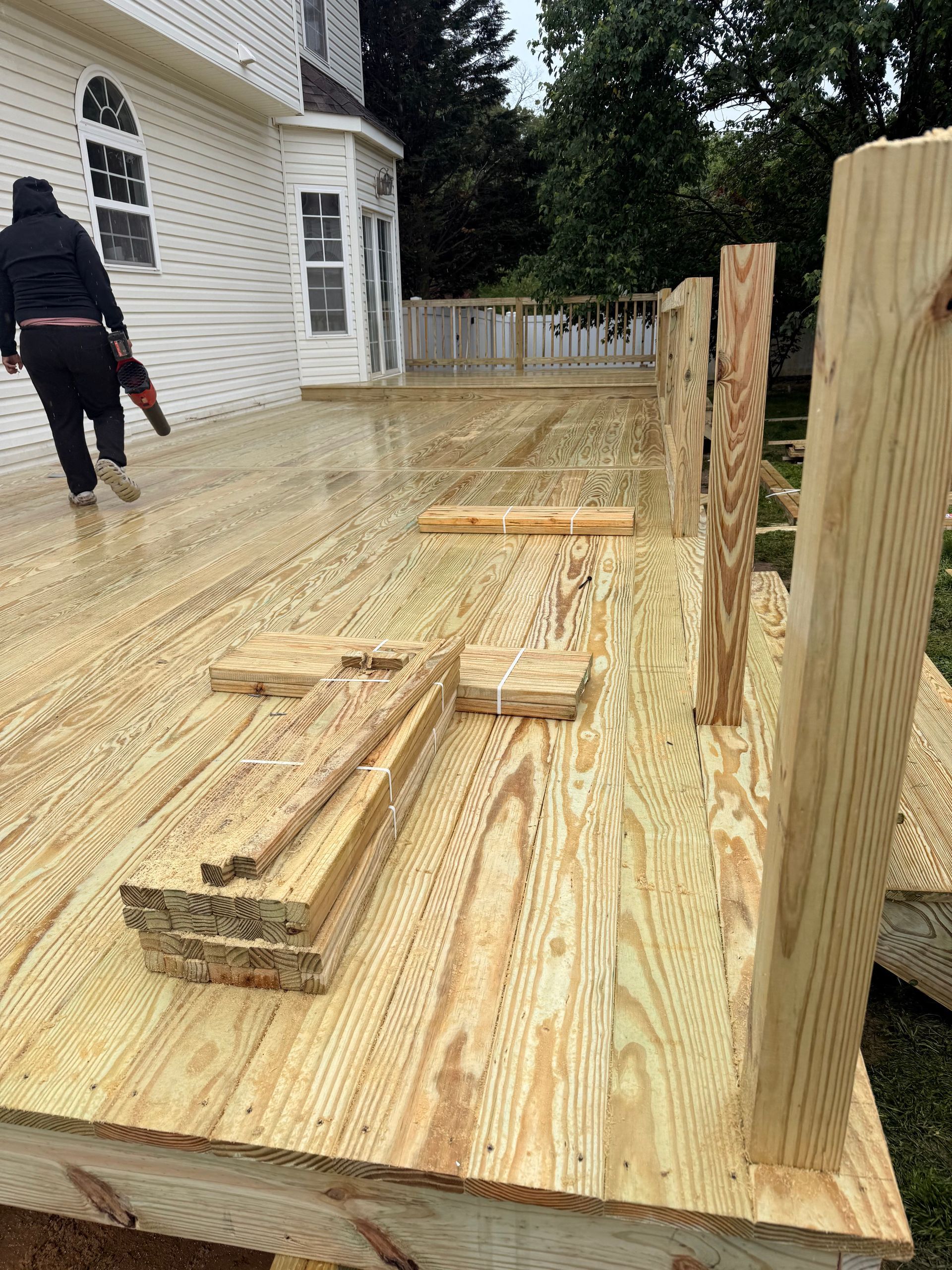 Wooden deck framing under construction beside a white house, with lumber and a worker on the left