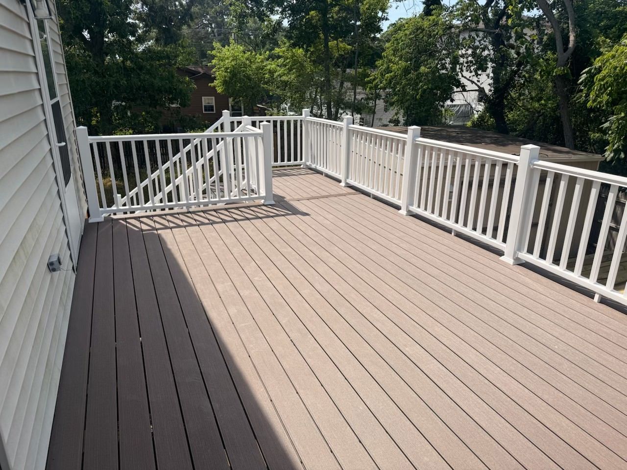 Spacious brown deck with white railings beside a house, overlooking trees and a fenced yard.