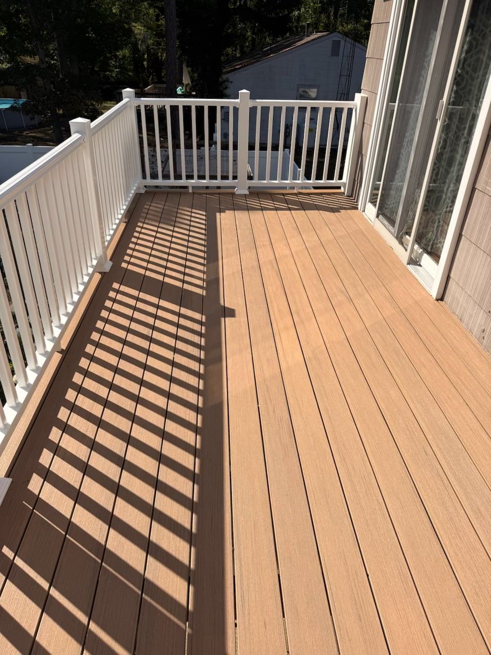 Sunlit wooden deck with white railing and long diagonal shadows, beside a house.