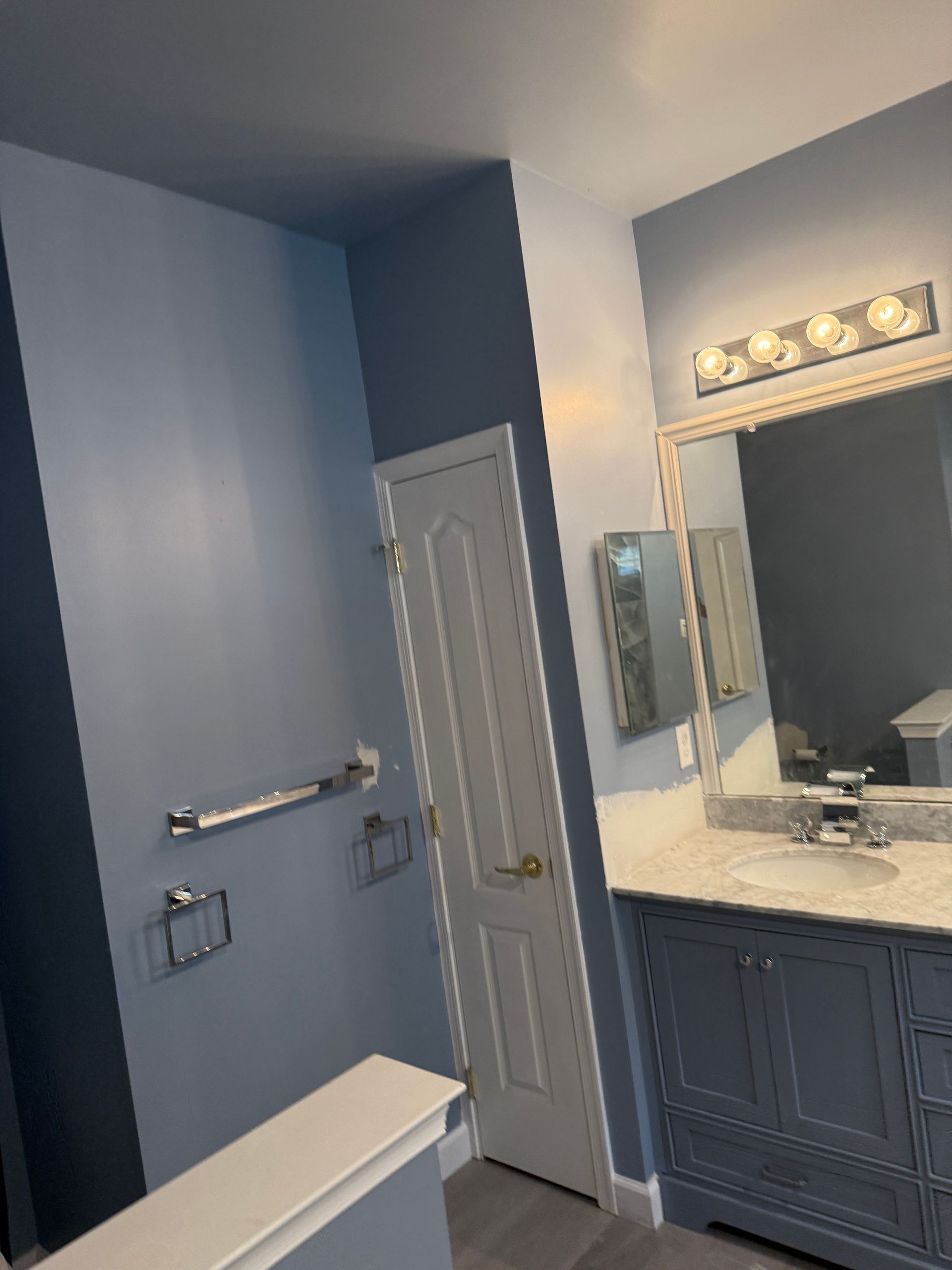 Blue bathroom with white door, wall towel bars, and vanity with mirror and lights.