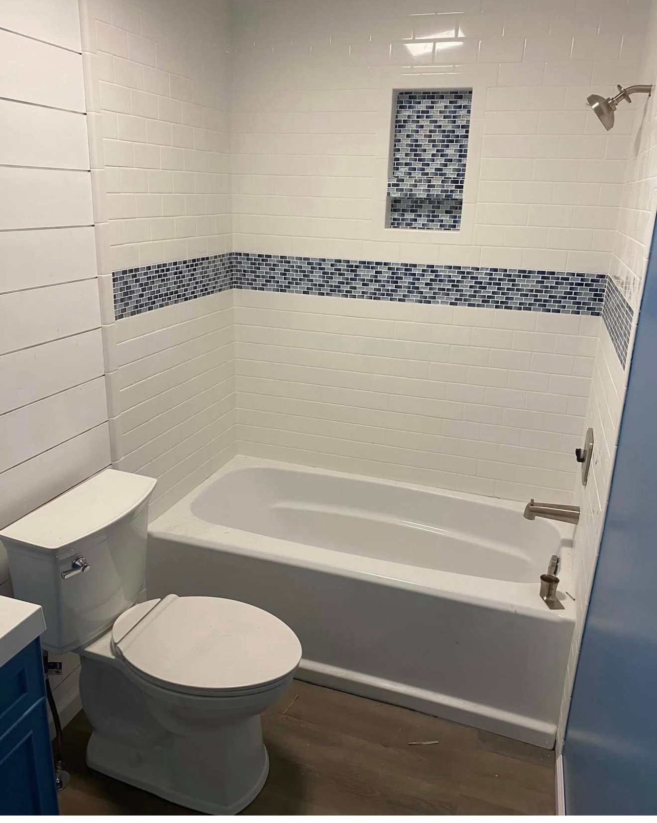 Small bathroom with white tub, toilet, blue tile accents, and wood-look floor