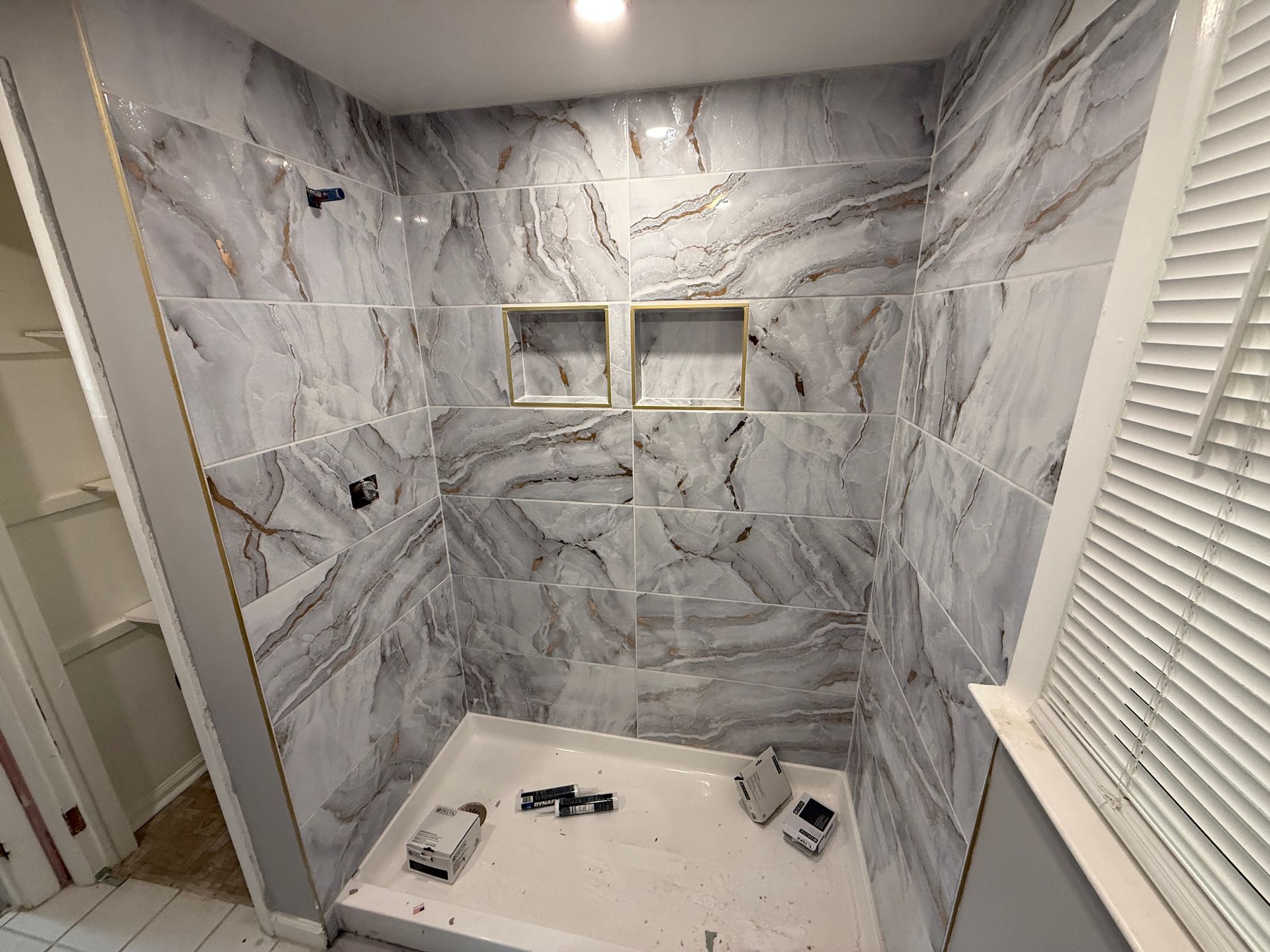 Marble-tiled shower under construction with a built-in bench, two niches, and a window with blinds.