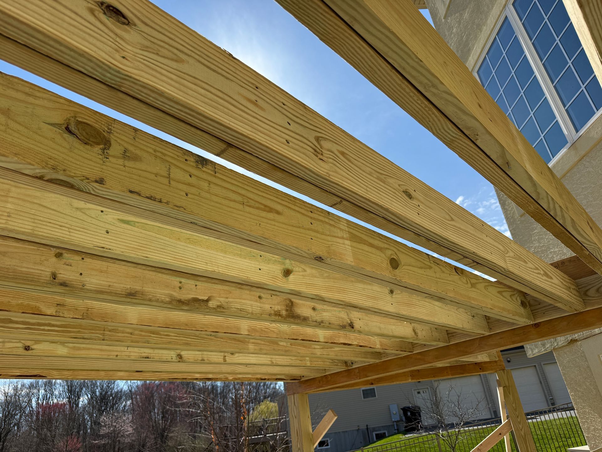 Wooden deck or pergola beams viewed from below beside a house under a blue sky.