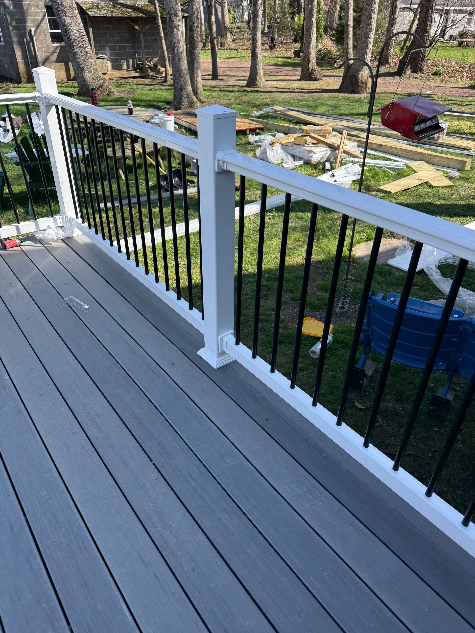 Gray deck railing with black balusters overlooking a wooded yard and scattered landscaping stones.