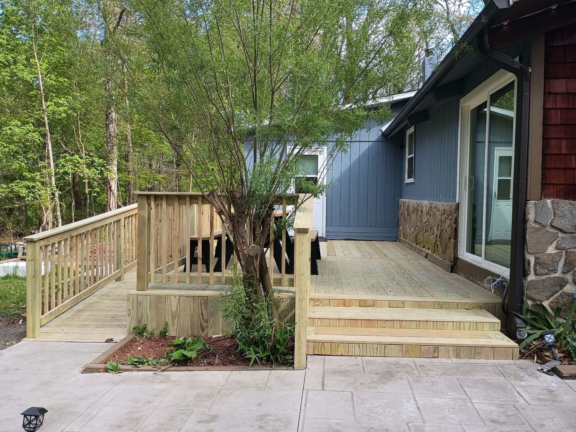 Wooden deck and wheelchair ramp beside a blue house, surrounded by trees and stone steps.