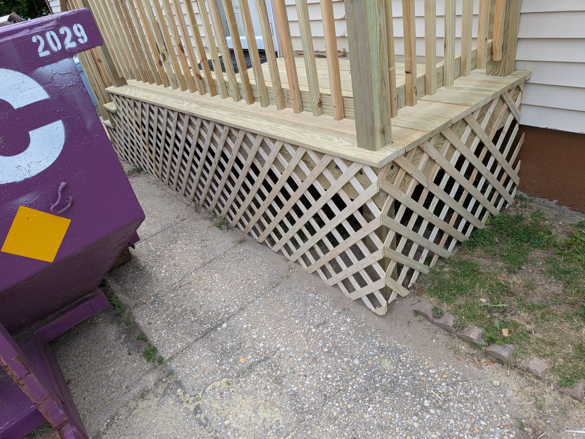 Concrete walkway beside a wooden porch, with a lattice skirt and a purple dumpster at left.