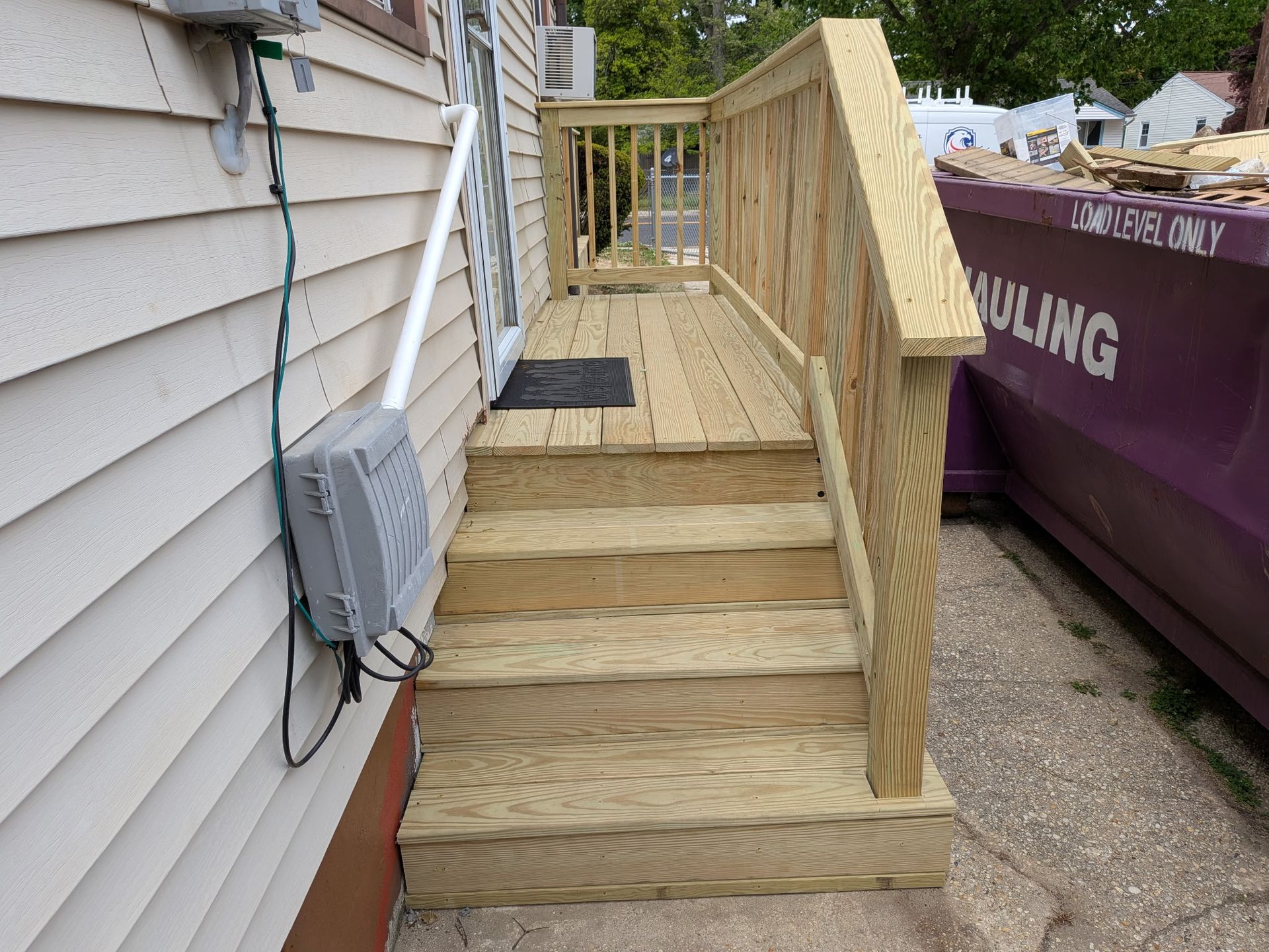 Wooden accessibility ramp with steps beside a house, bordered by rails and a dumpster on the right.
