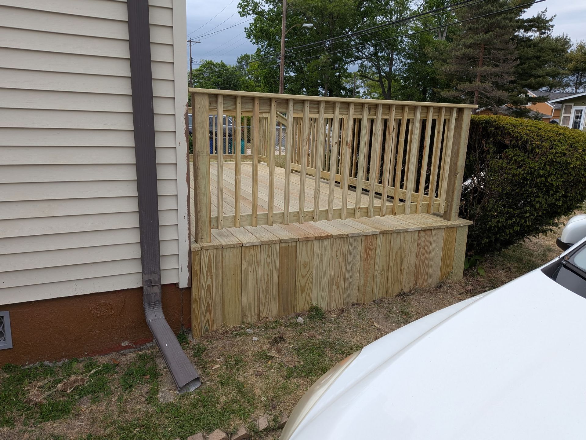 Wooden deck with railing beside a house, next to a white car in a residential yard.