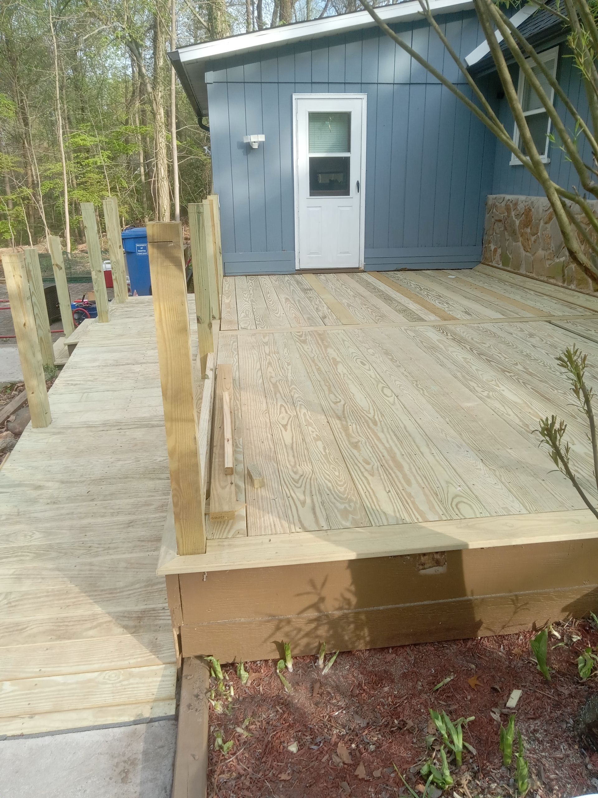 Wooden ramp leads to a small blue building with a white door in a wooded outdoor area.