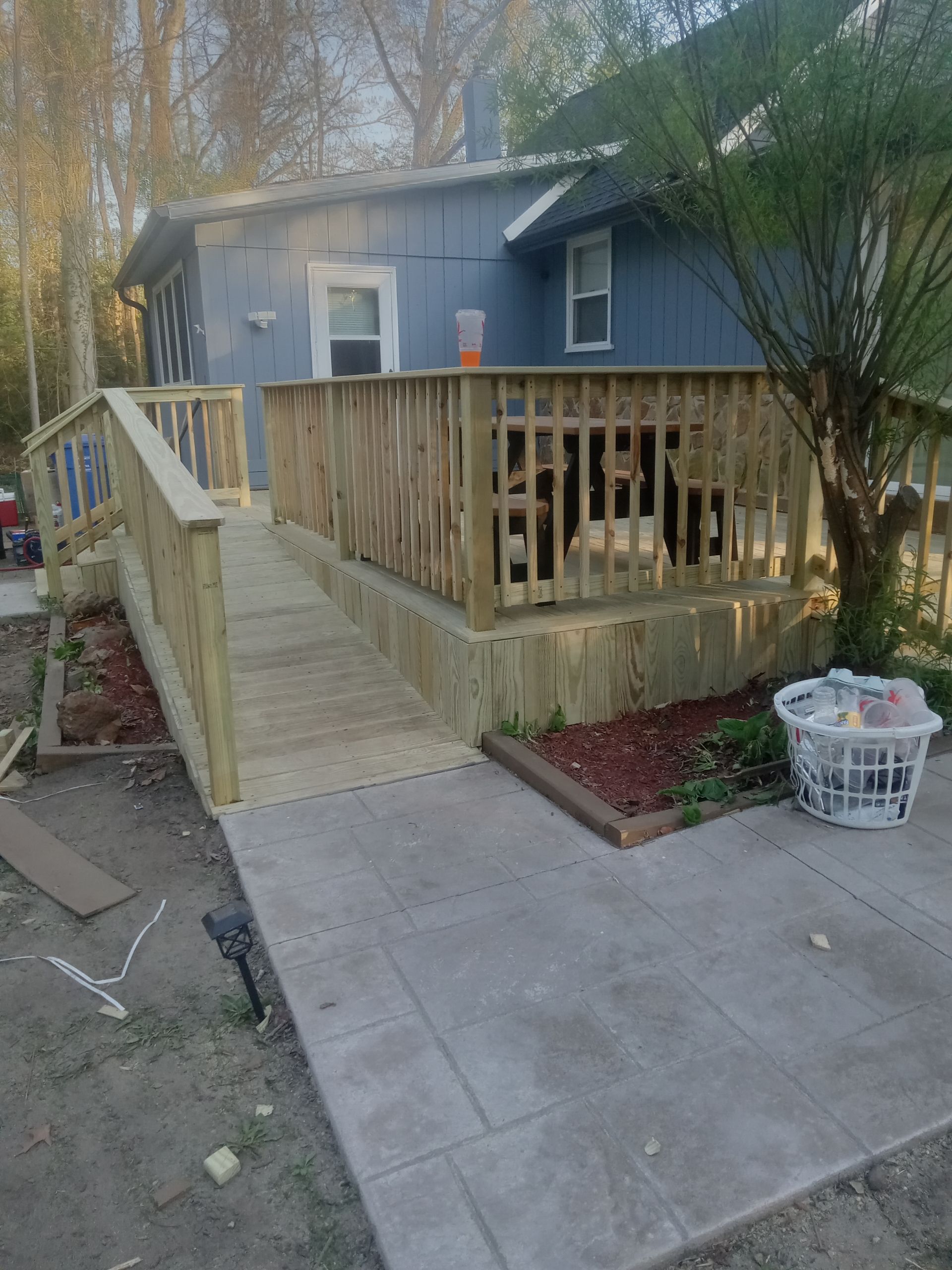 Front porch with wooden ramp and railings leading to a blue house, with a driveway and laundry basket nearby