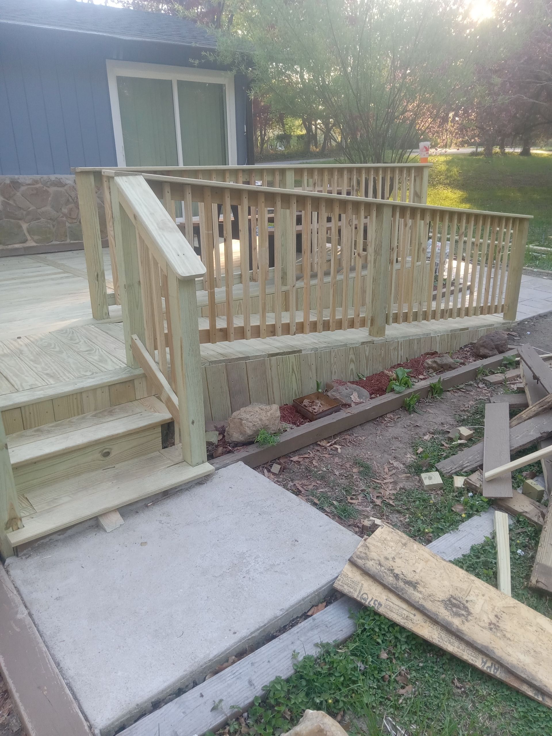 Wooden ramp and stairs leading to a porch beside a blue house, with concrete path and landscaping.