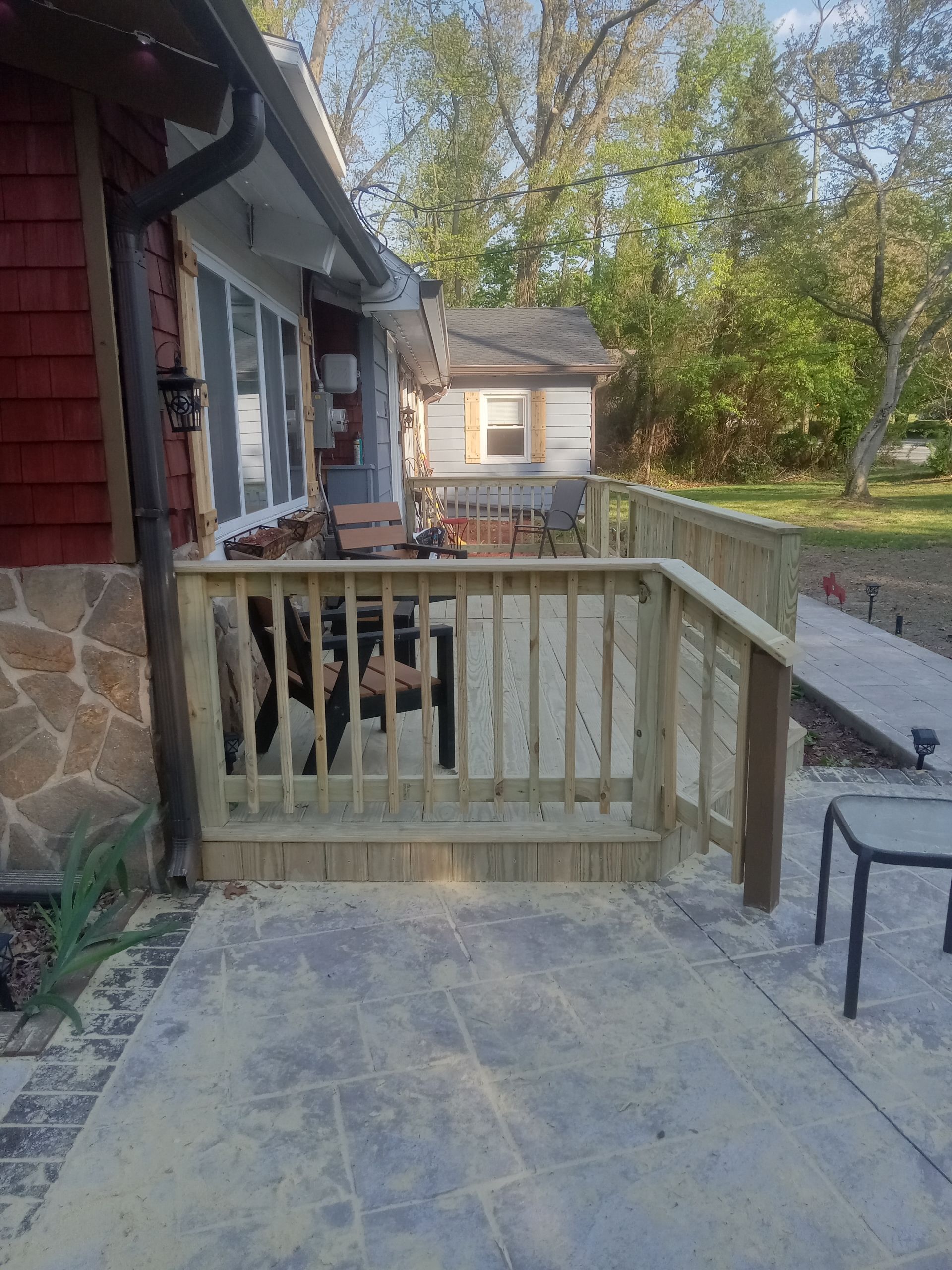Front porch with wooden railing, stone patio, and steps beside a house in a leafy yard