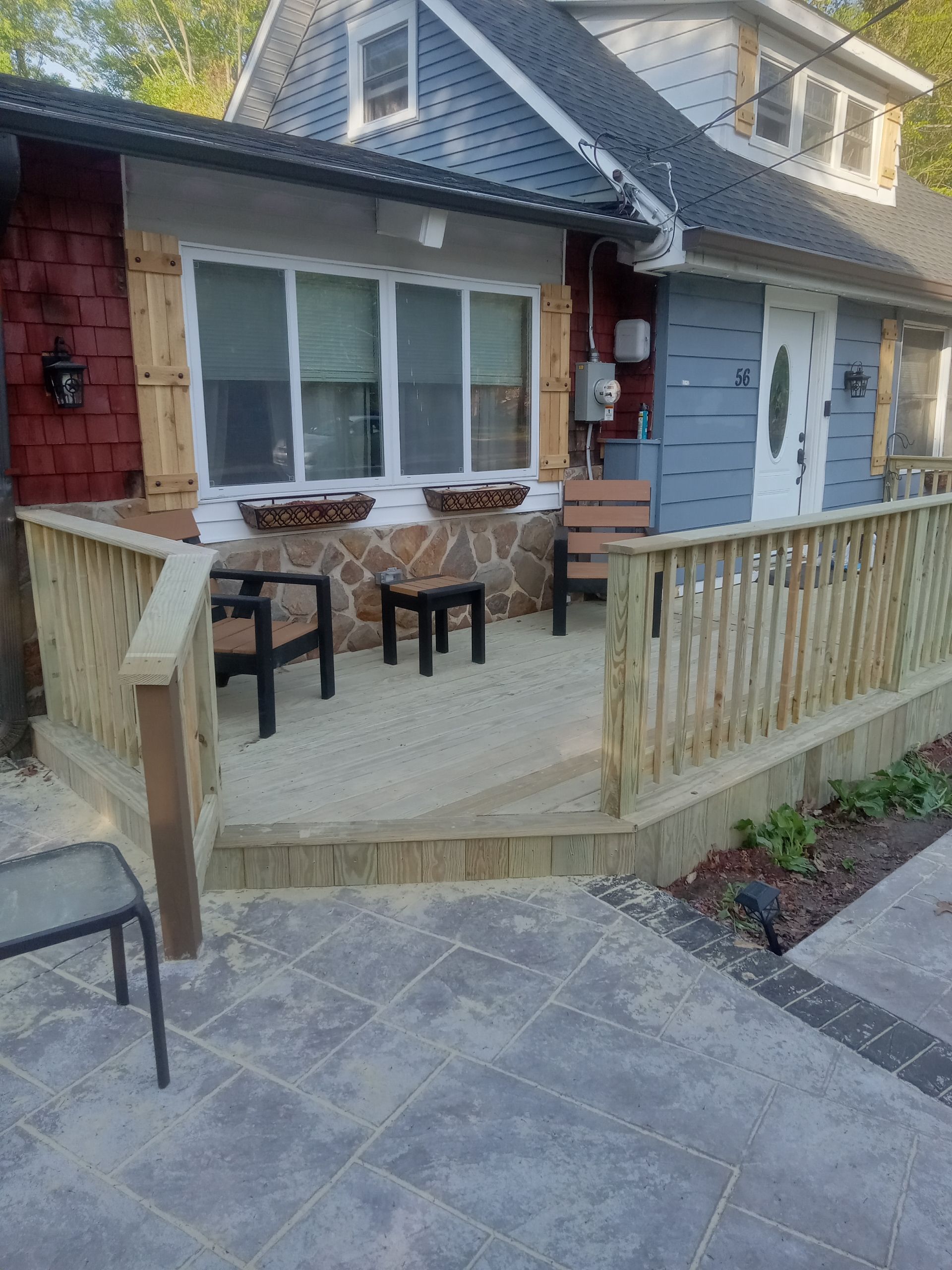 Front porch with stone pavers, wooden railing, chairs, and a house entrance with windows and blue siding