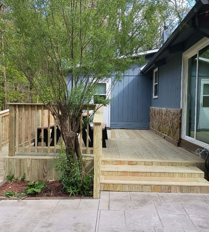 Wooden deck with steps beside a blue house, bordered by a fence and trees.