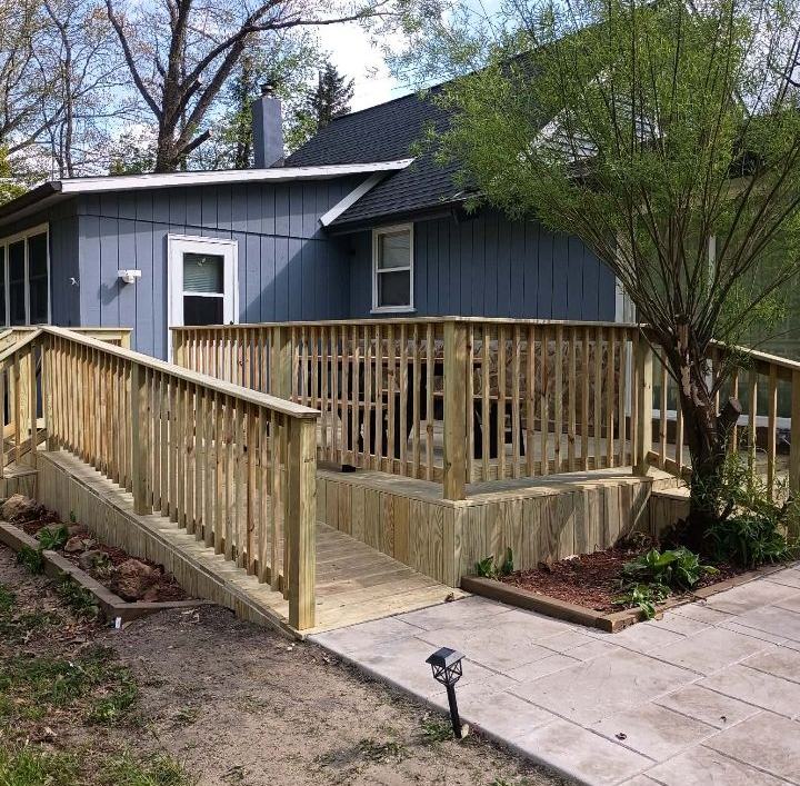 Wooden wheelchair ramp and deck beside a blue house with a concrete walkway and trees