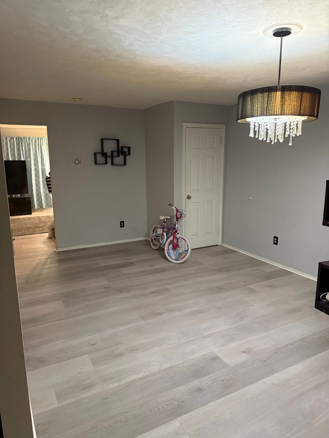 Interior room with light wood-look flooring, gray walls, and a chandelier. A child's pink bicycle stands inside.
