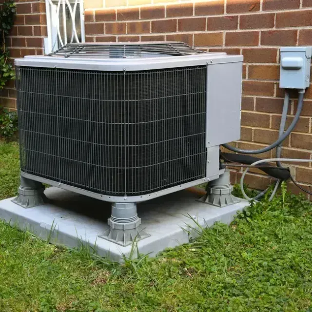 Air conditioning unit on a concrete pad, against a brick wall, resting on elevated supports.
