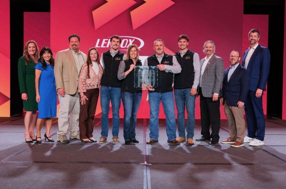 Ten people stand in a line on a stage with a red Lennox backdrop, posing while two central members hold an award plaque.
