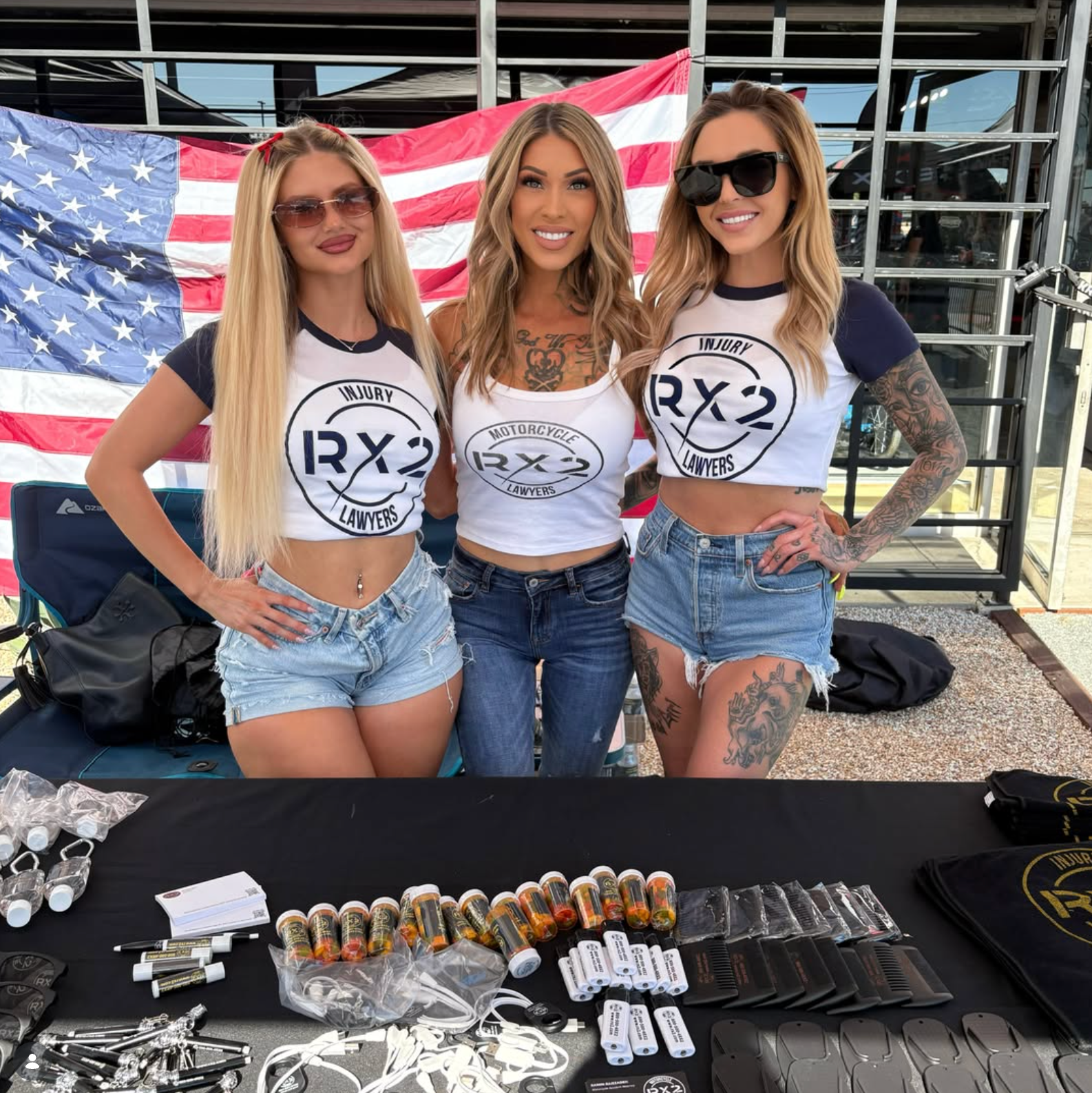 Three women in matching RX3 shirts and denim shorts standing behind a table of merchandise with an American flag backdrop