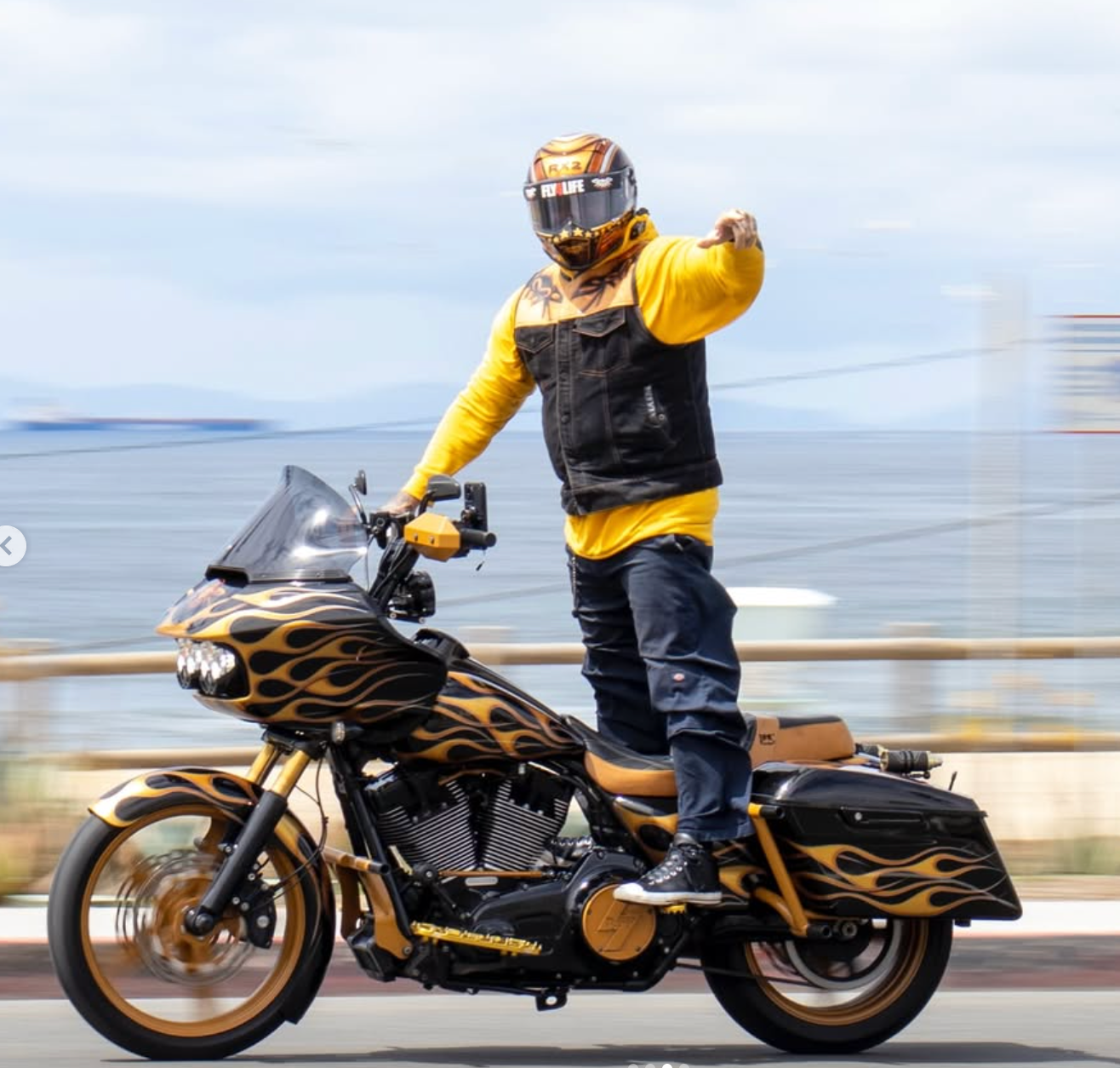Motorcyclist in yellow and black gear riding a flame-decaled bike, waving on a blurred road by the water
