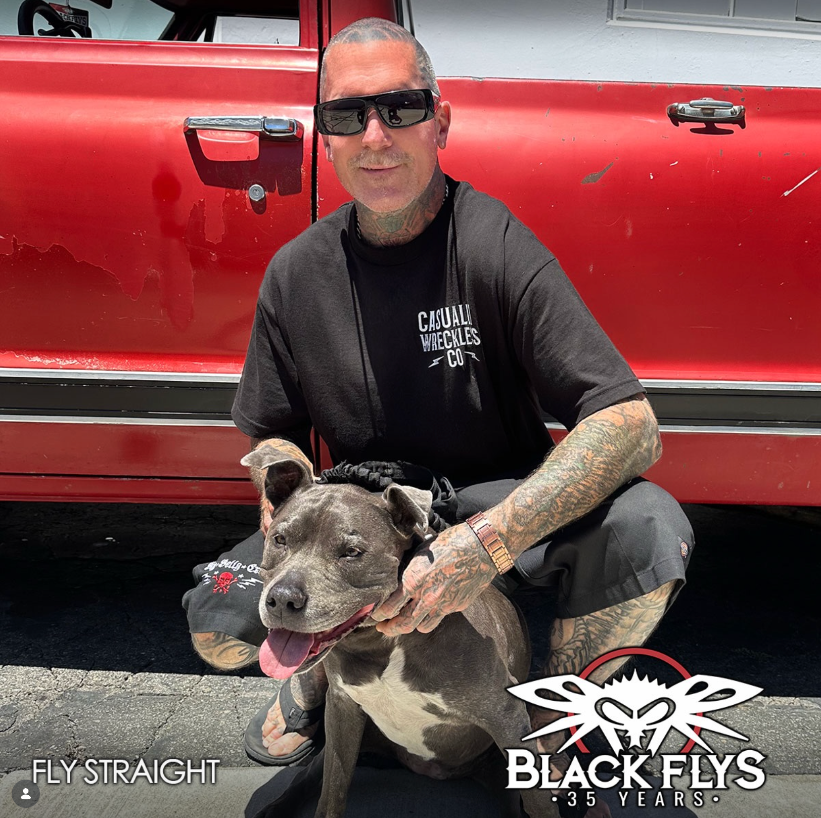Man crouching beside a gray pit bull in front of a red vehicle, both wearing black clothing.
