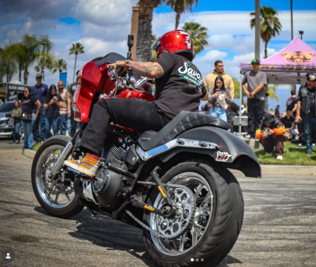 Motorcyclist on a red bike performing a stunt at a sunny outdoor event with palm trees and spectators.