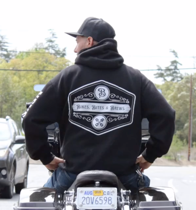Person on a motorcycle wearing a black hoodie and cap, viewed from behind on a tree-lined street.