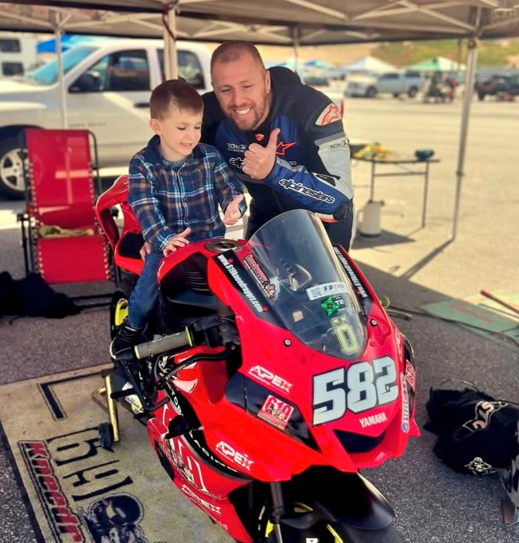 Man and child posing on a red racing motorcycle under a canopy, number 582 visible.