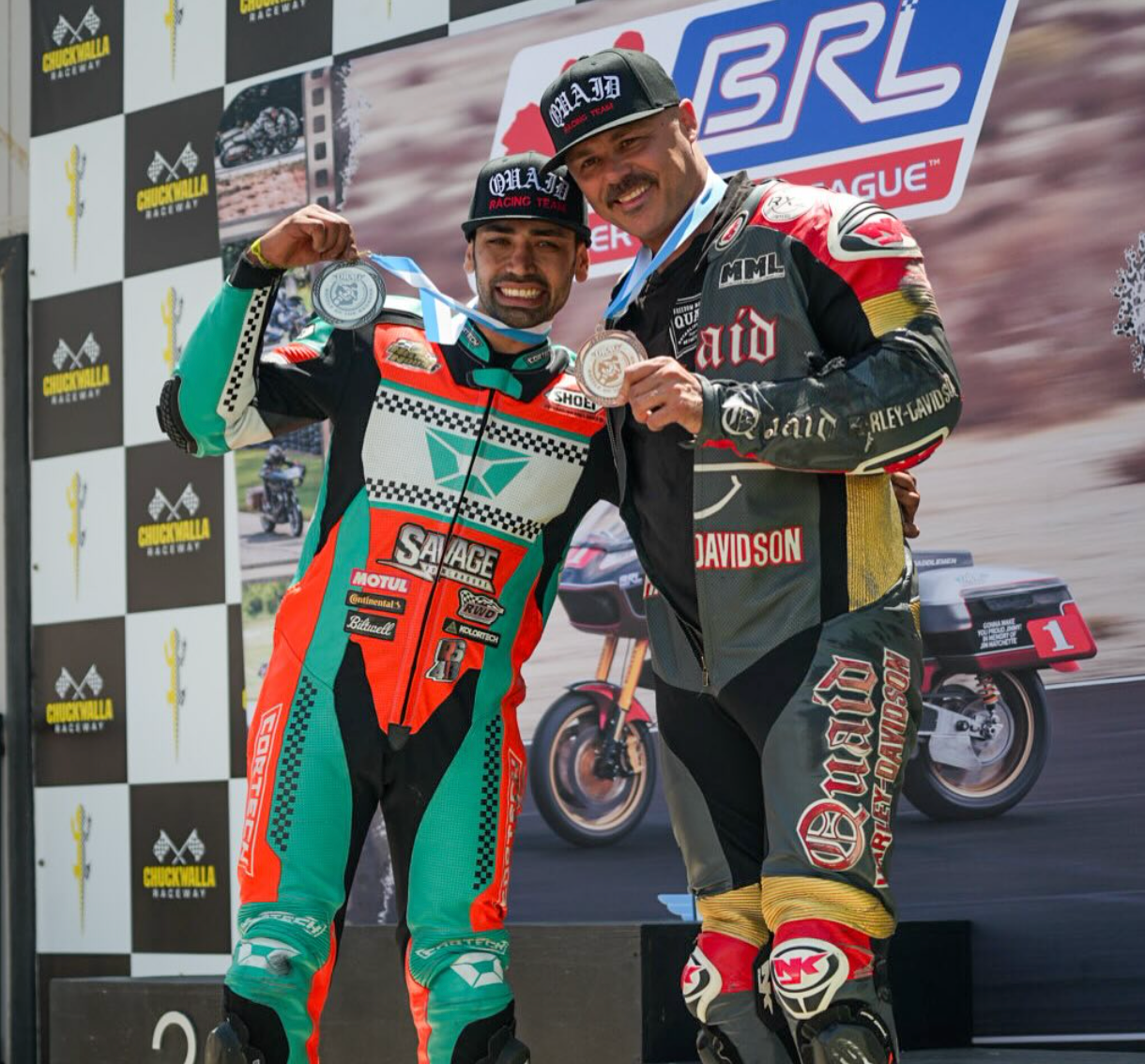 Two race car drivers in colorful suits pose with trophies on a podium at a GRL event.