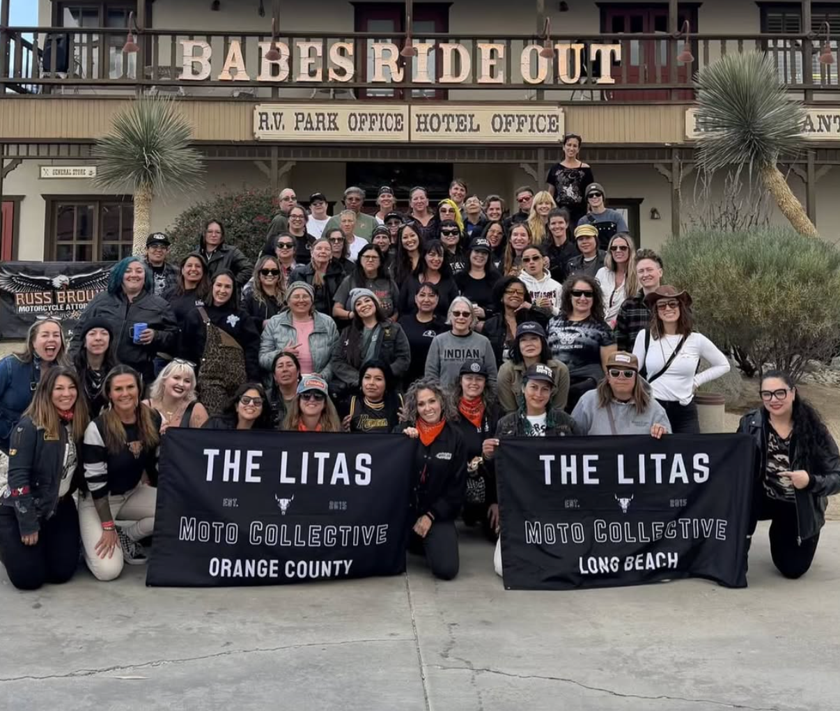 Large group posing outside a theater, holding two black banners reading “The Litas,” in front of “Babes Ride Out” sign