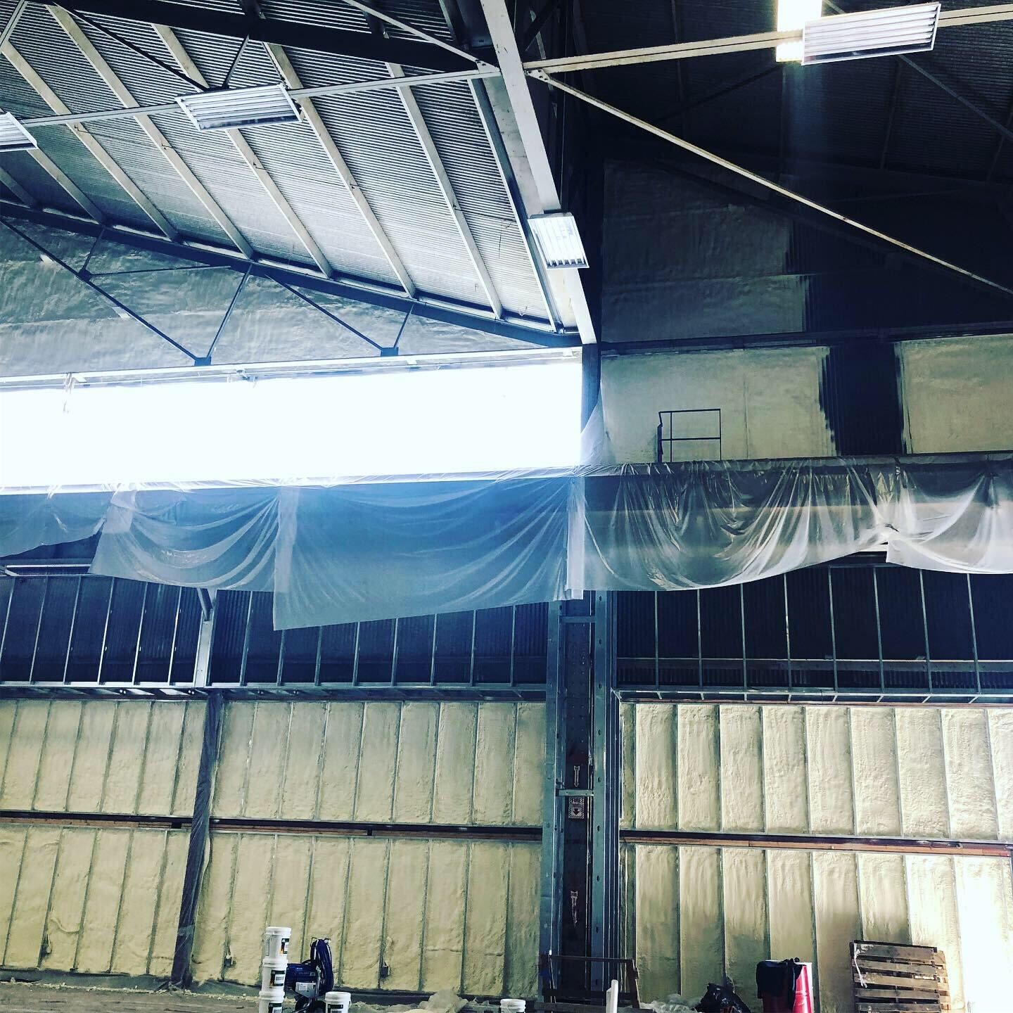 Interior of a warehouse with metal roof beams, hanging white netting, and stacked insulation panels.