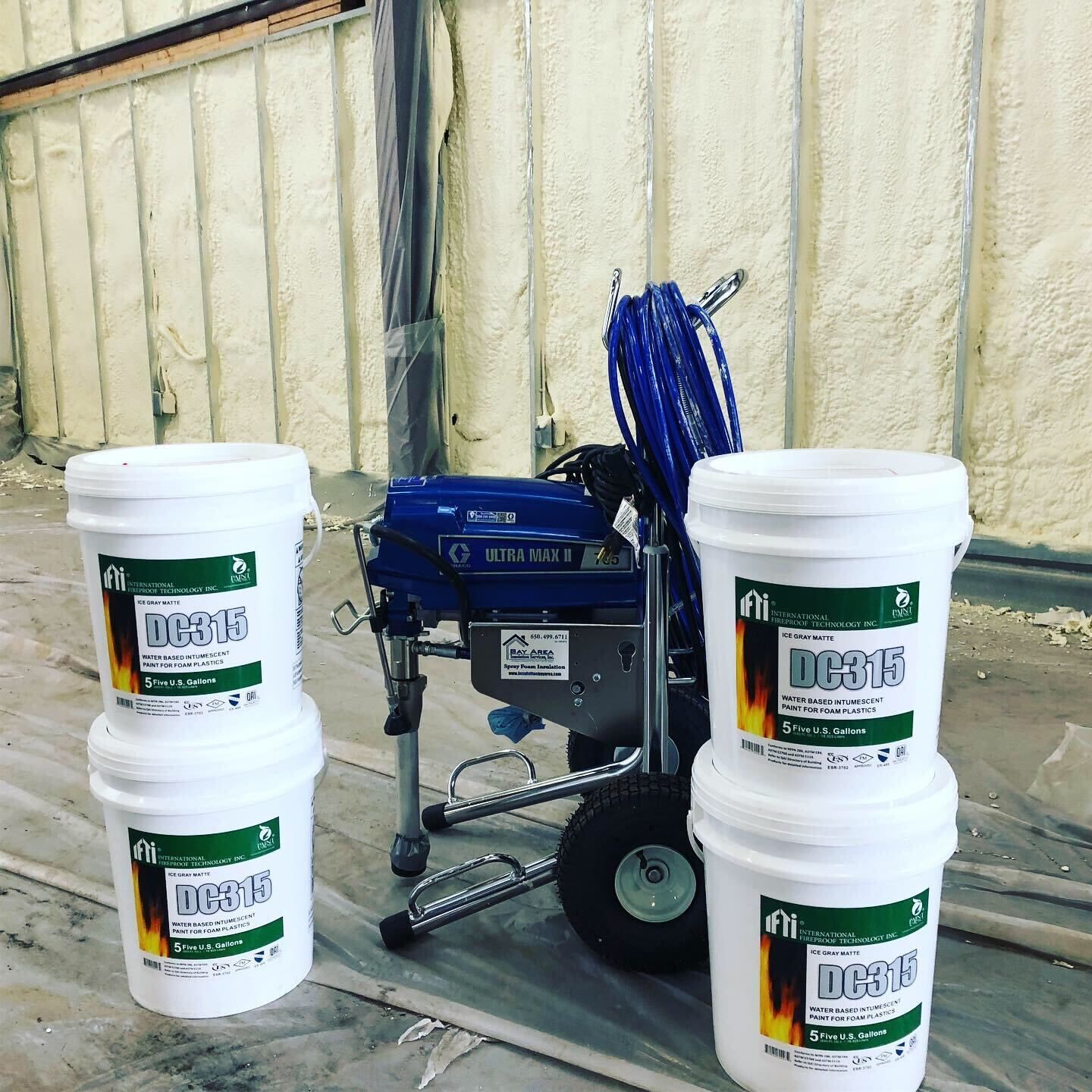 Blue paint sprayer beside four white paint buckets in an indoor workspace