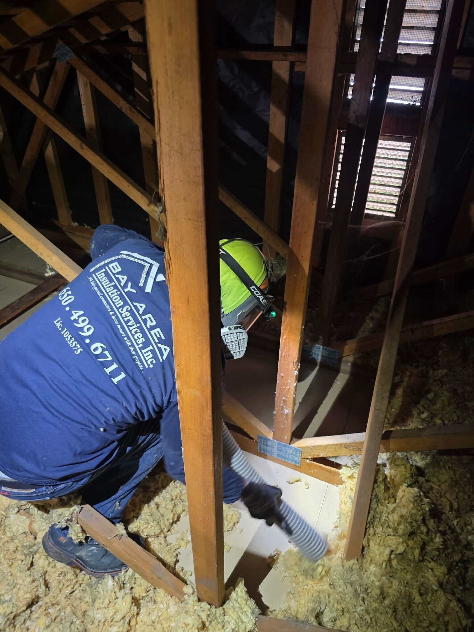 Worker in a blue shirt and hard hat crouches in an attic with exposed beams and insulation.