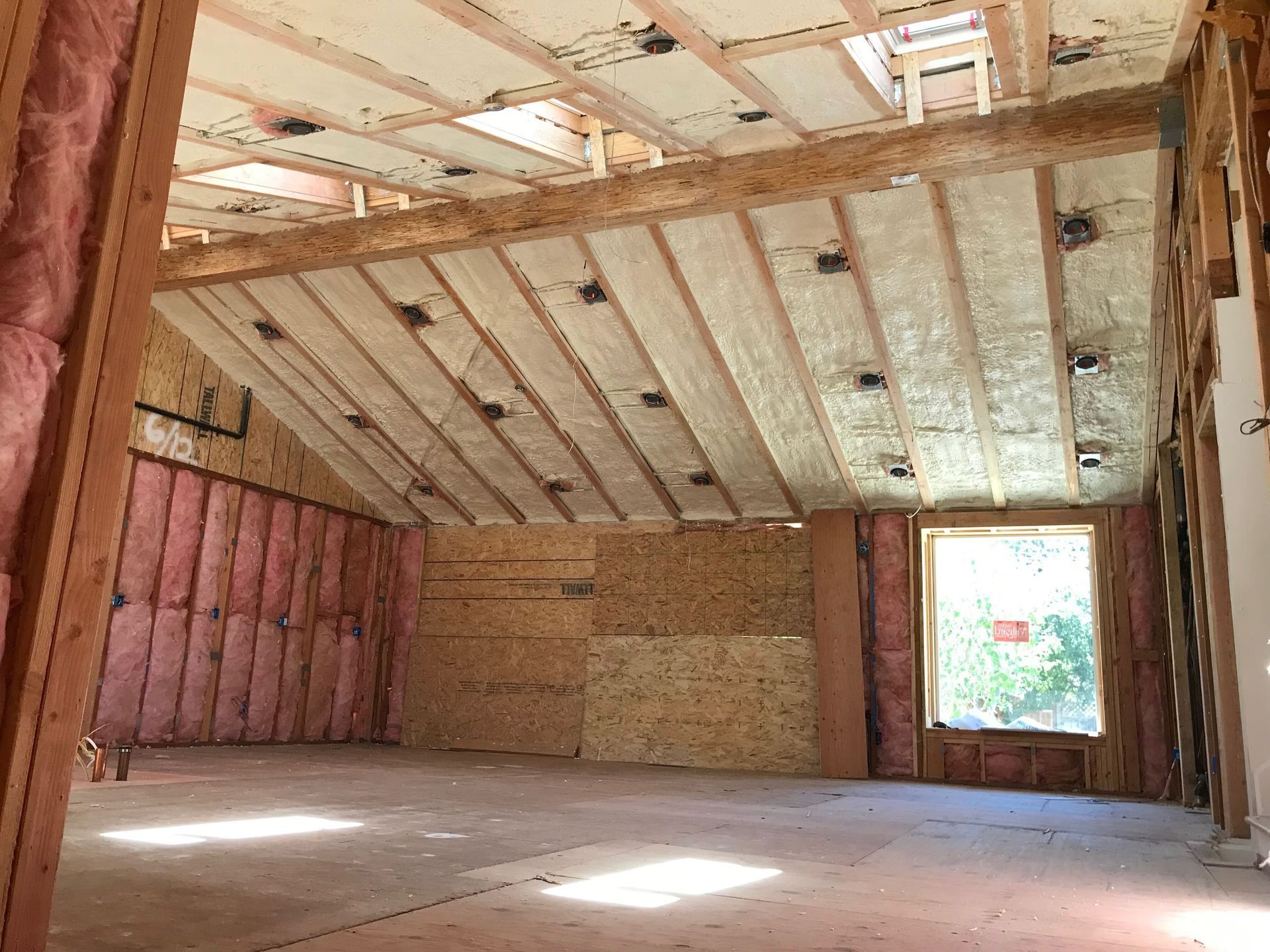 Empty unfinished room with exposed insulation, angled ceiling beams, and sunlight from a window.