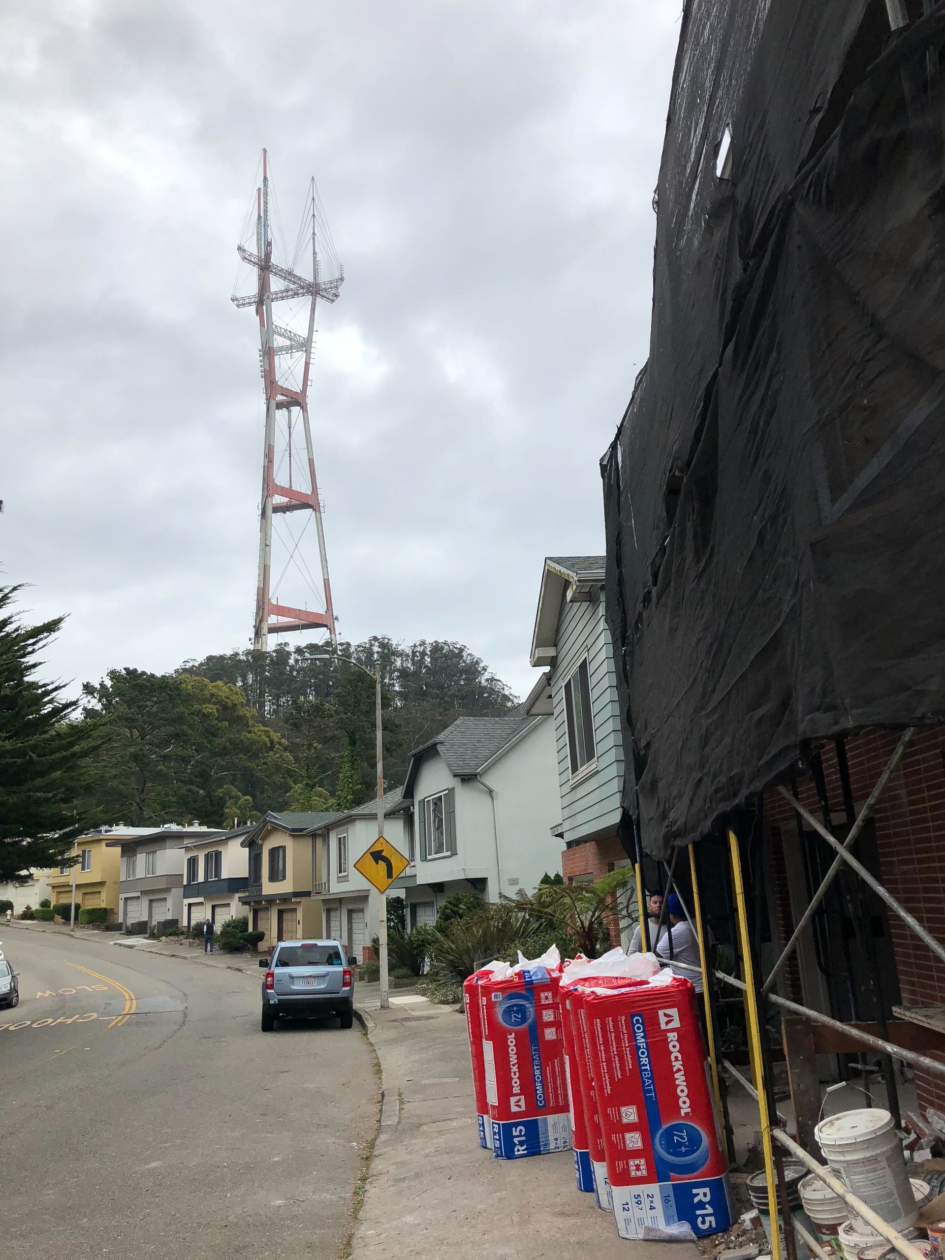 Steep street with parked cars, red bins, and a tall radio tower under cloudy sky