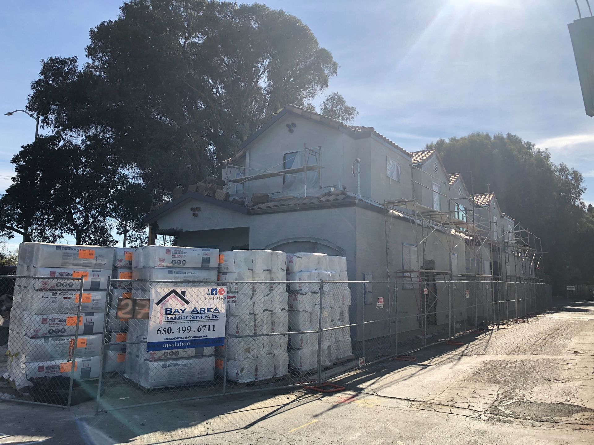 Sunlit construction site with stacked materials, scaffolding, and a large house under renovation
