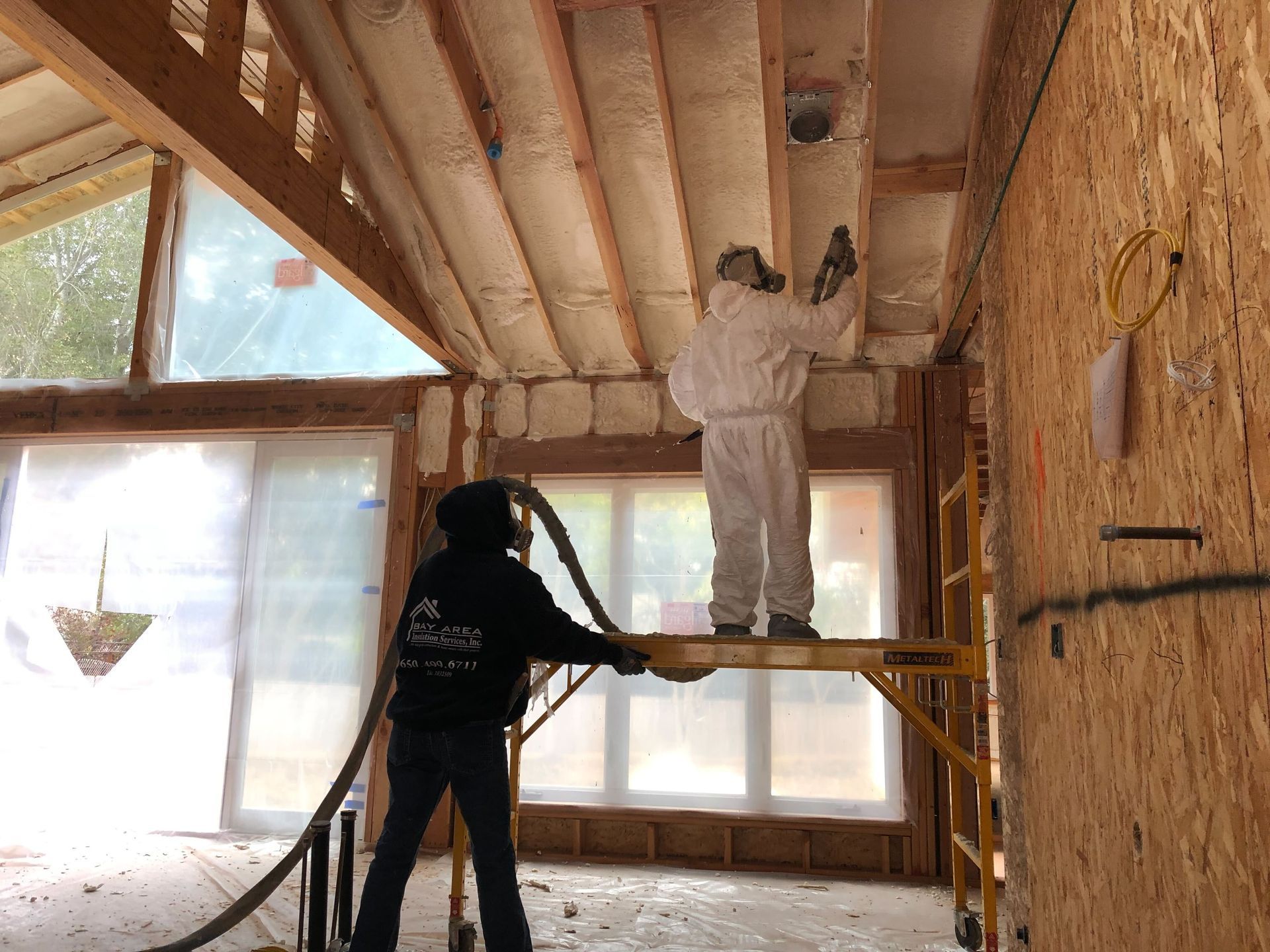 Two workers insulating a cabin ceiling, one on a scaffold and one spraying foam into rafters