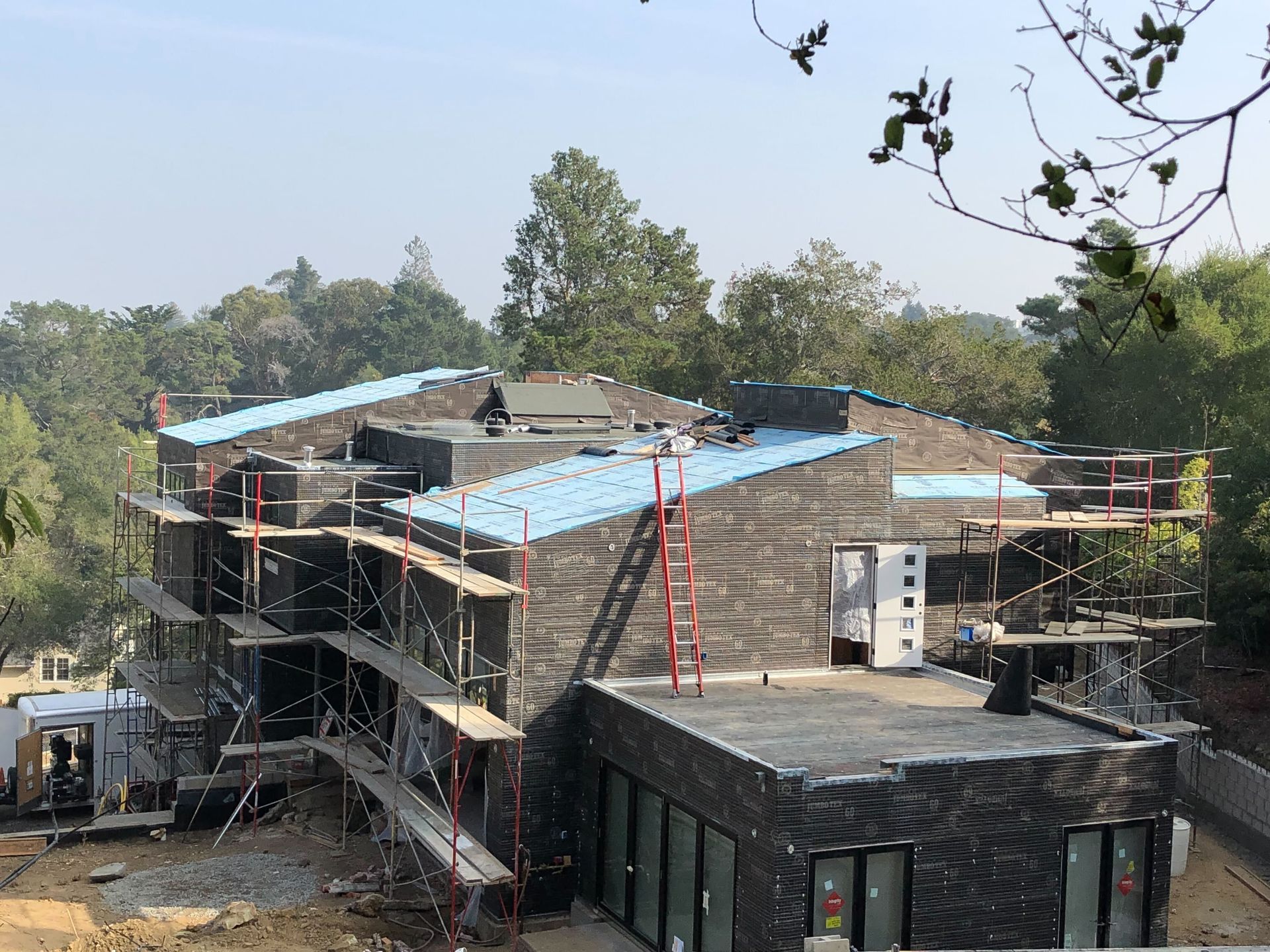 Two-story building under construction with blue tarp roofing and scaffolding on a wooded site