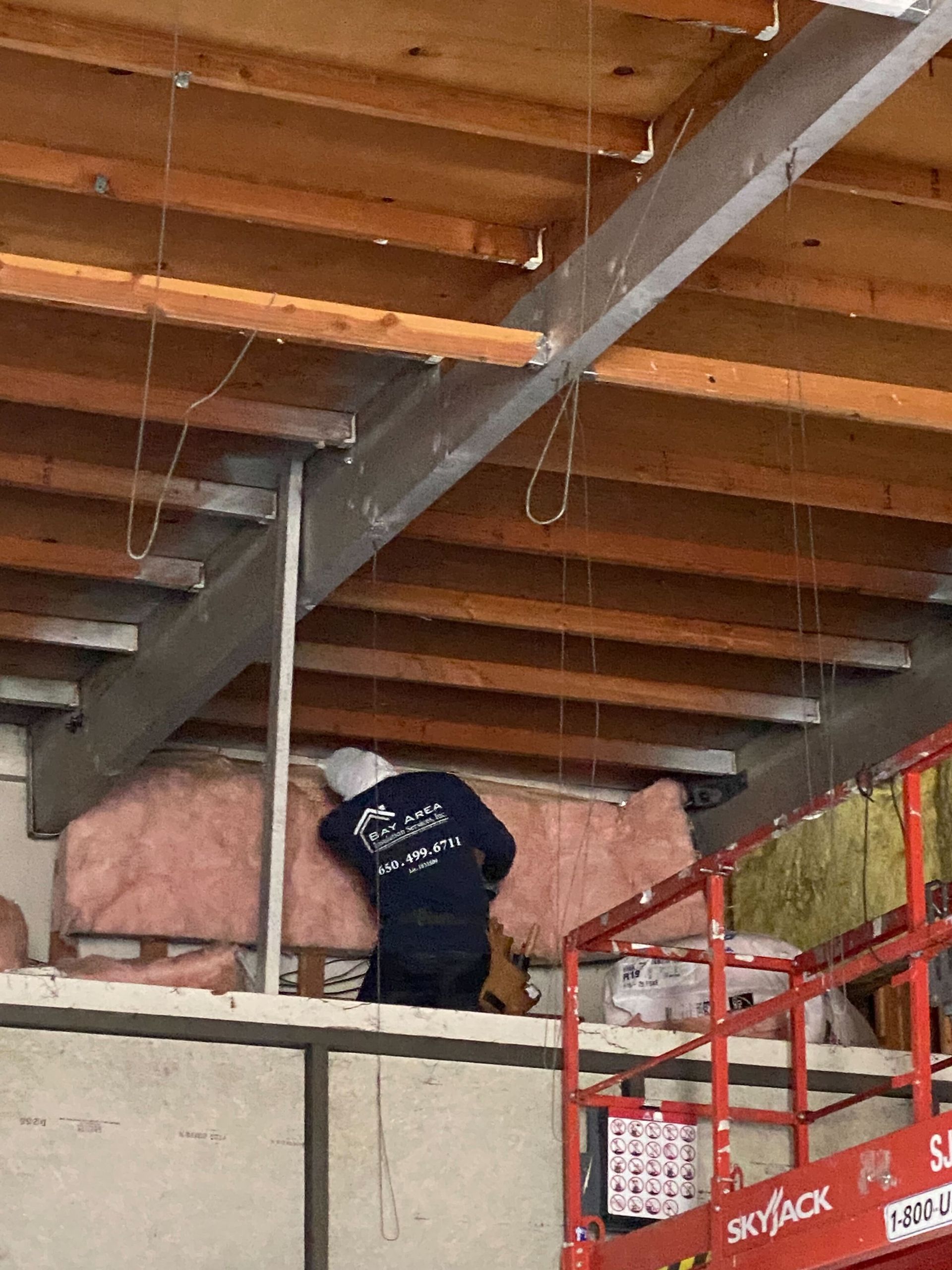 Worker on a red lift in an unfinished building, handling insulation near exposed beams.