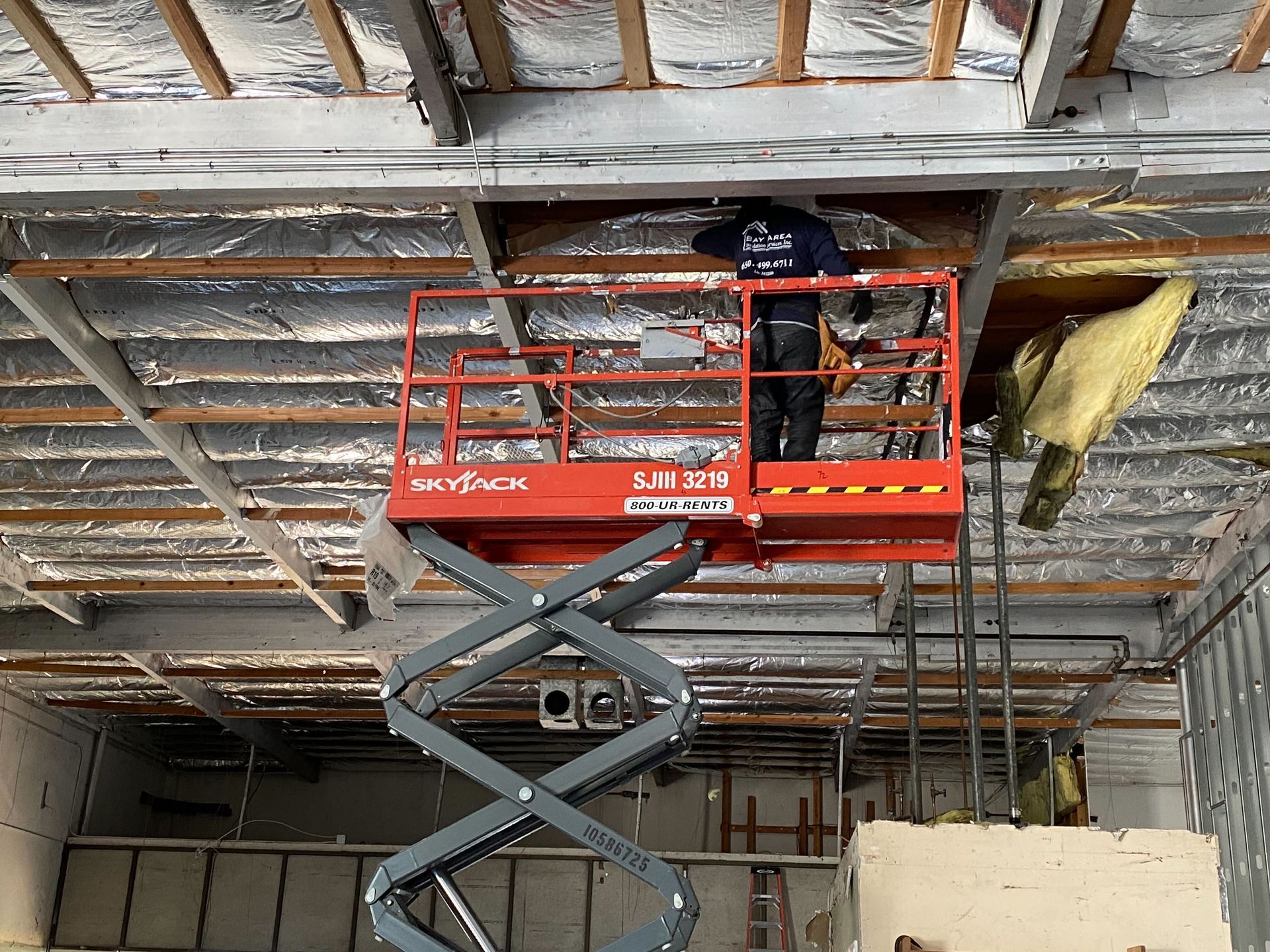 Worker on red lift repairing a damaged ceiling with exposed insulation and beams.