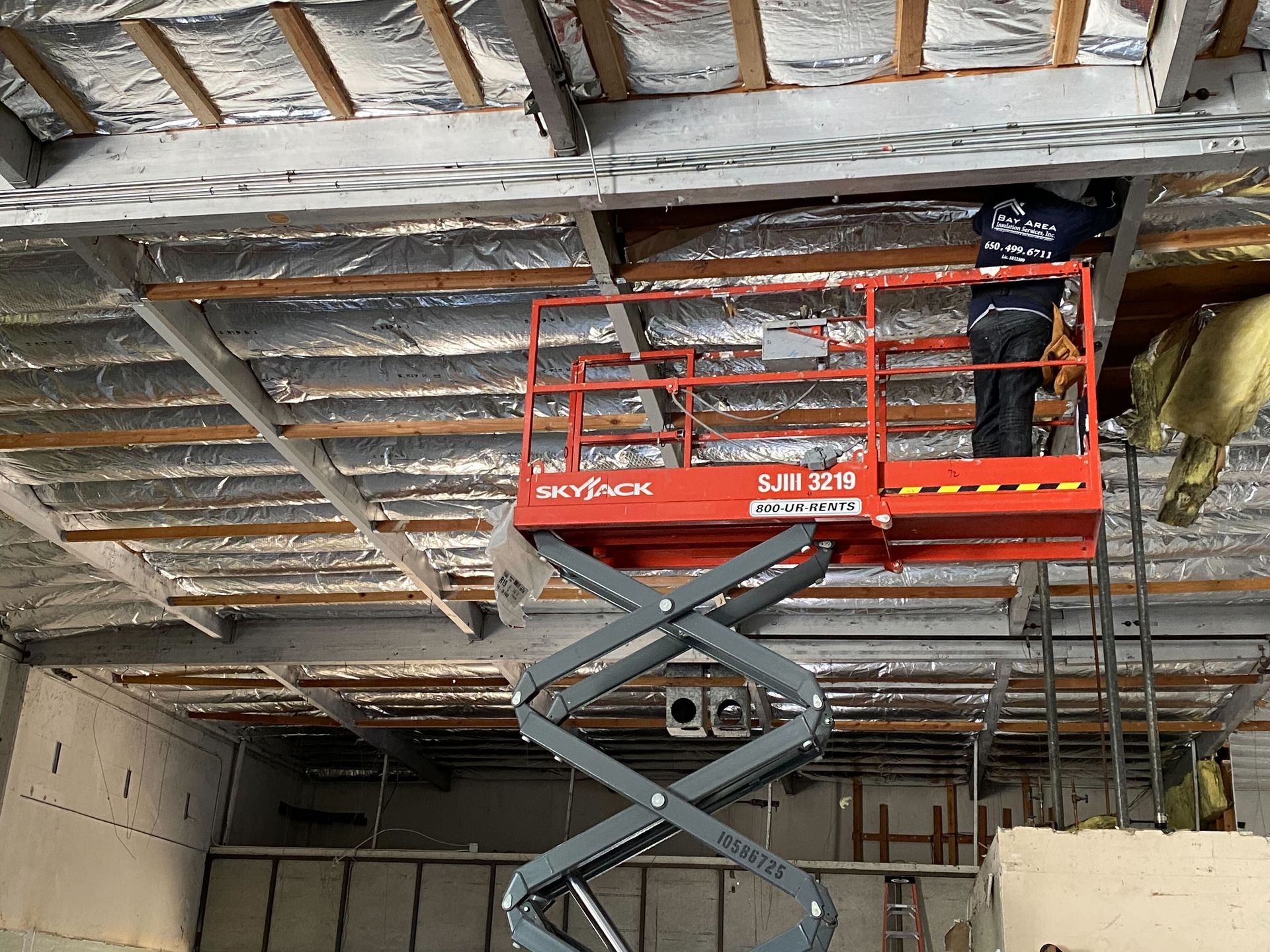 Worker on a red scissor lift installing ceiling insulation in an unfinished interior.