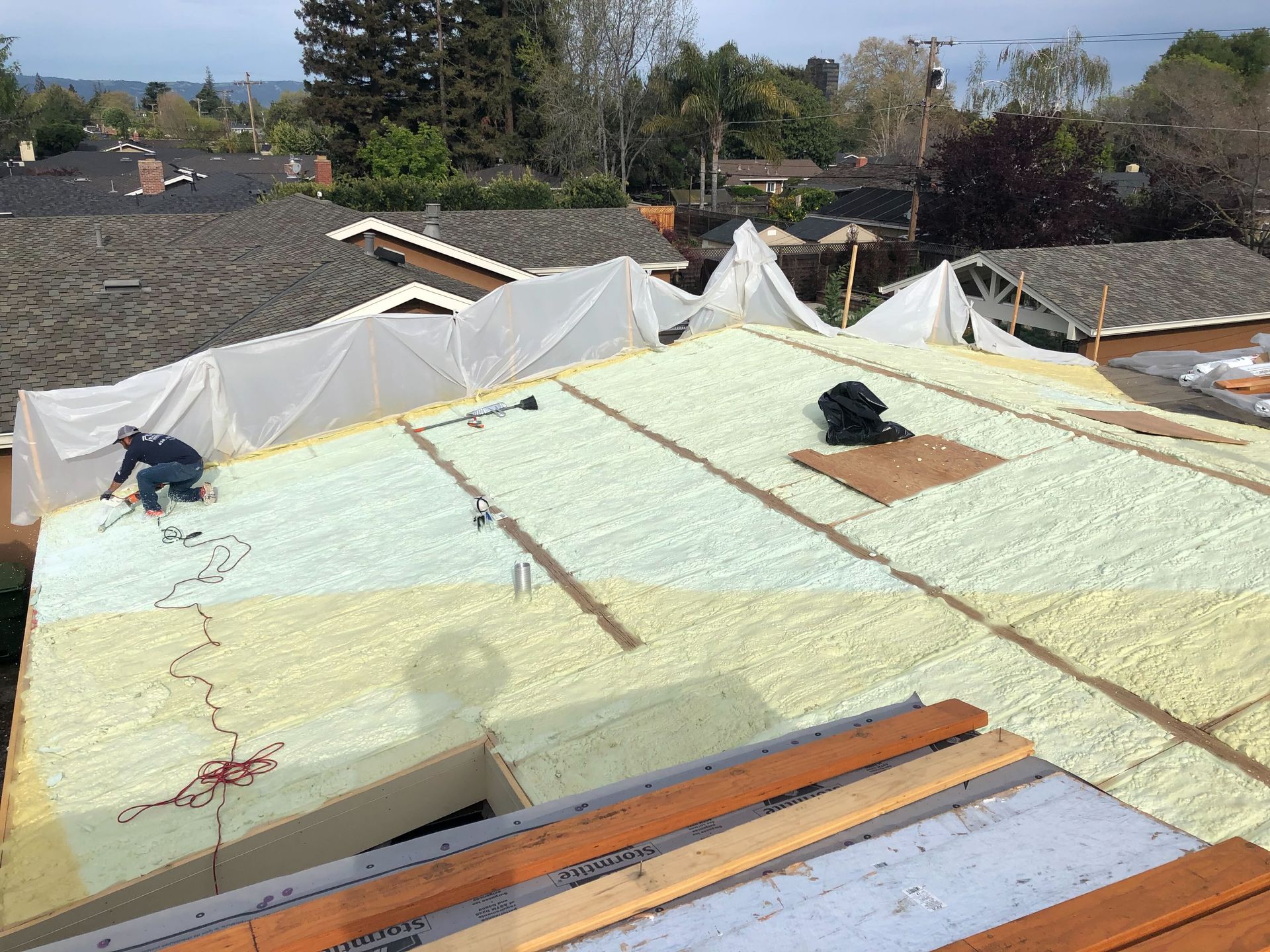Roof under construction with yellow insulation foam, white tarp, and rooftop vents in a residential area