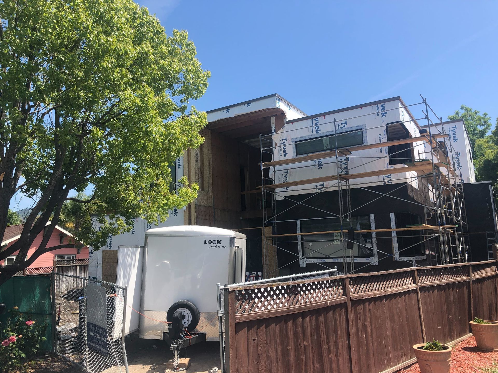 House under construction behind scaffolding and fence, with a white trailer and leafy tree in front