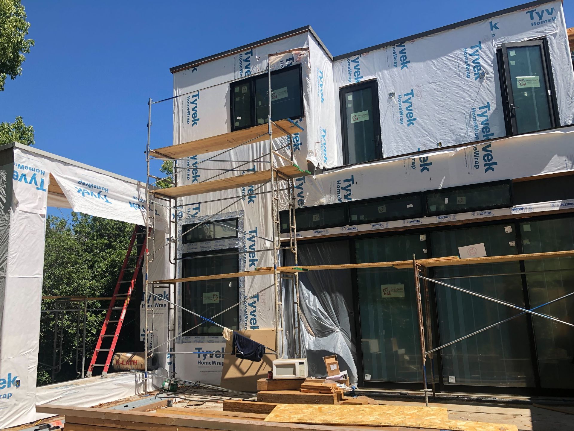 Two-story house under construction with scaffolding, ladders, and wrapped exterior in daylight