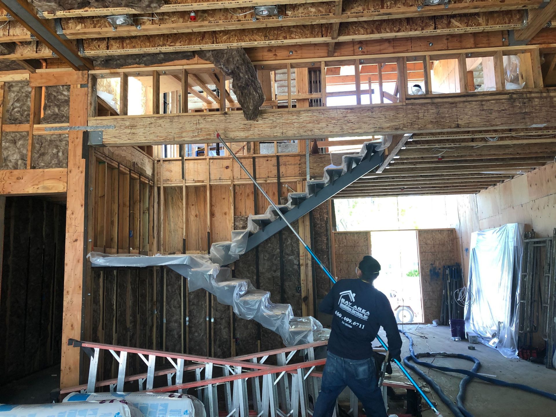 Construction worker on a ladder in a wood-framed house, inspecting a staircase under construction.