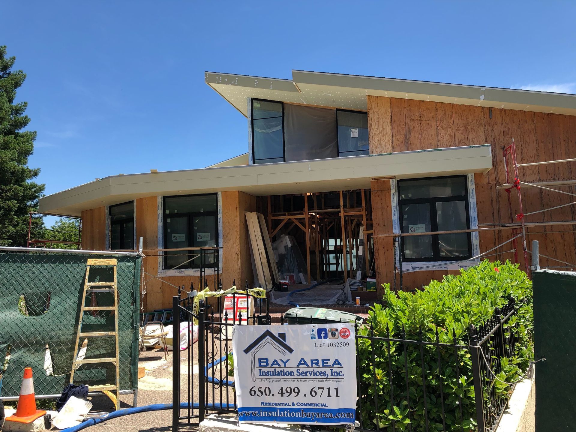 House under construction with scaffolding, ladder, and front yard shrubs under a blue sky