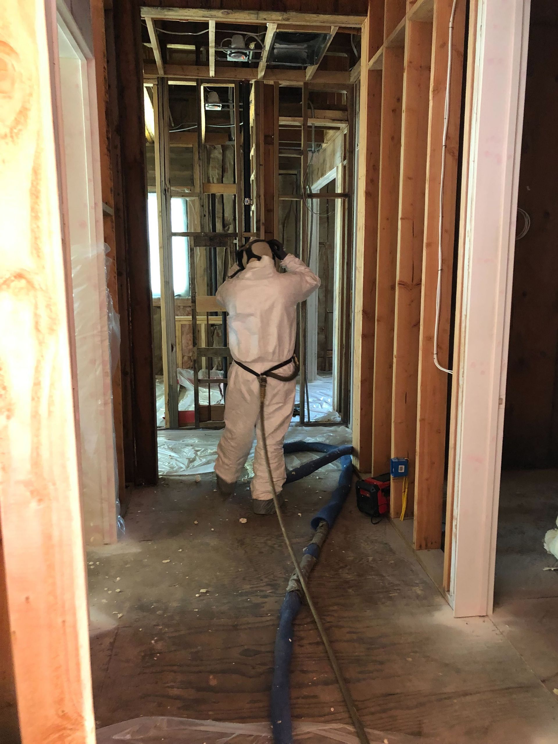 Person spraying foam insulation inside an unfinished hallway with exposed framing.