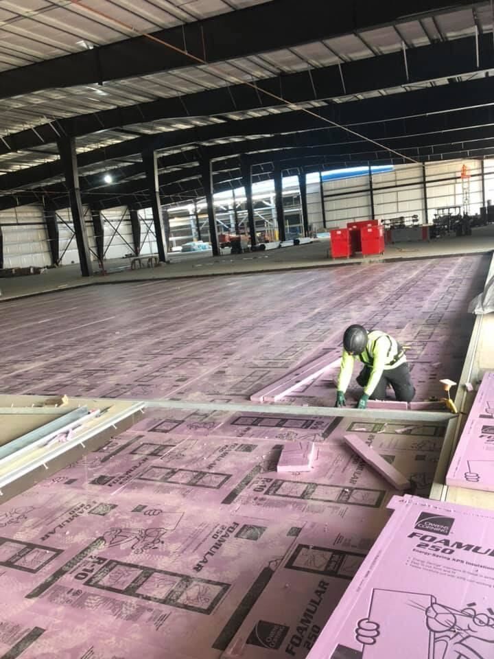 Worker installing pink insulation panels in a large unfinished warehouse interior.